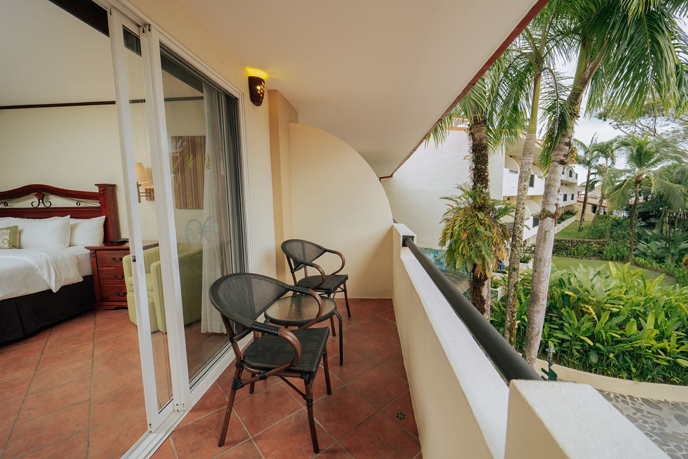 A balcony with a bed, two chairs, and a view of a garden with palm trees.