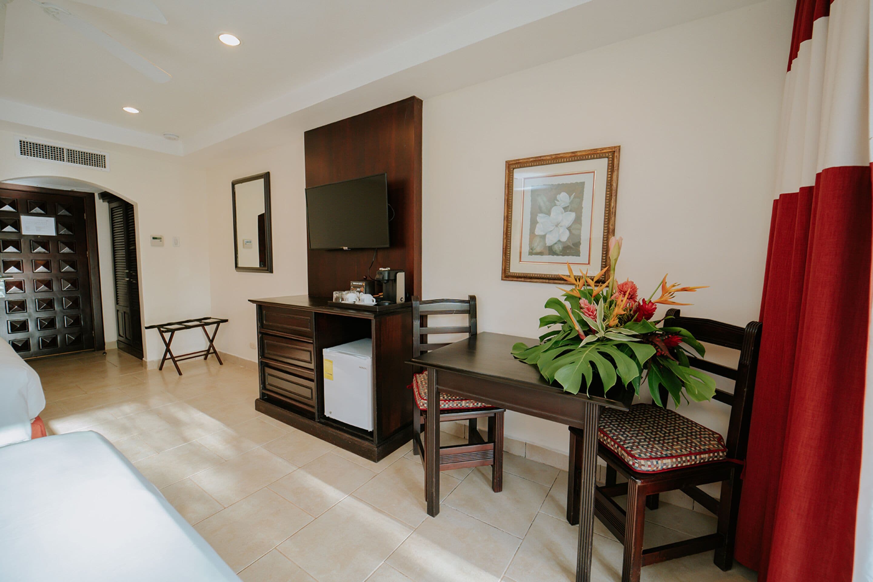 A hotel room with a dining area, a TV mounted on the wall, a framed picture, and a plant on the table.