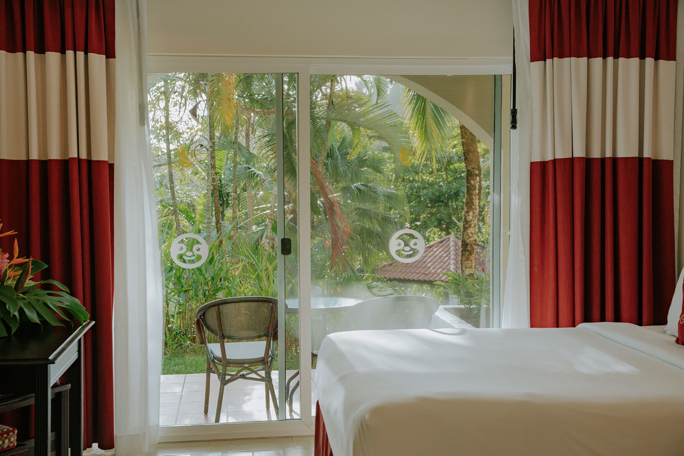 Une chambre comprenant un lit, une chaise et une porte-fenêtre coulissante donnant sur un balcon avec vue sur la verdure et un toit recouvert de tuiles.