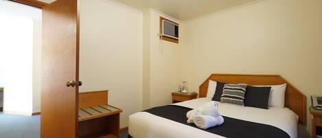 A hotel room with a bed, a bedside table with a lamp, a small window, and a door.