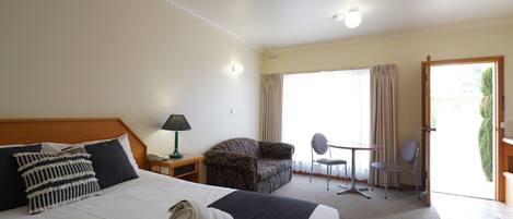 Une chambre d’hôtel avec un grand lit, un petit bureau avec une lampe, un canapé, un fauteuil et une vue sur l’extérieur par une porte.