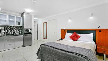 A modern hotel room with a kitchen area, a bed with a grey bedspread and red pillow, and a wooden nightstand.
