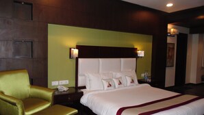 Quarto de hotel com uma cama grande, uma poltrona dourada, uma mesa de cabeceira de madeira com abajur e uma parede verde decorativa.