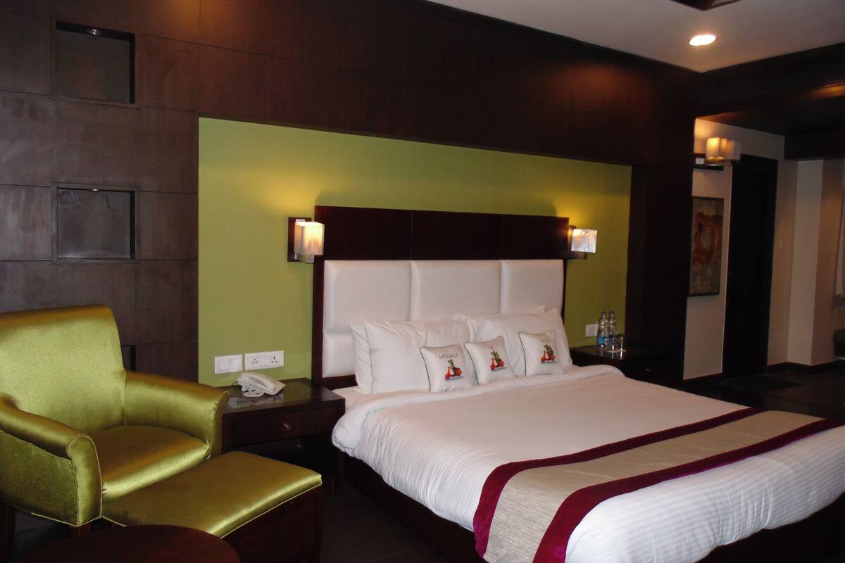 A hotel room with a large bed, a gold armchair, a wooden nightstand with a lamp, and a green accent wall.