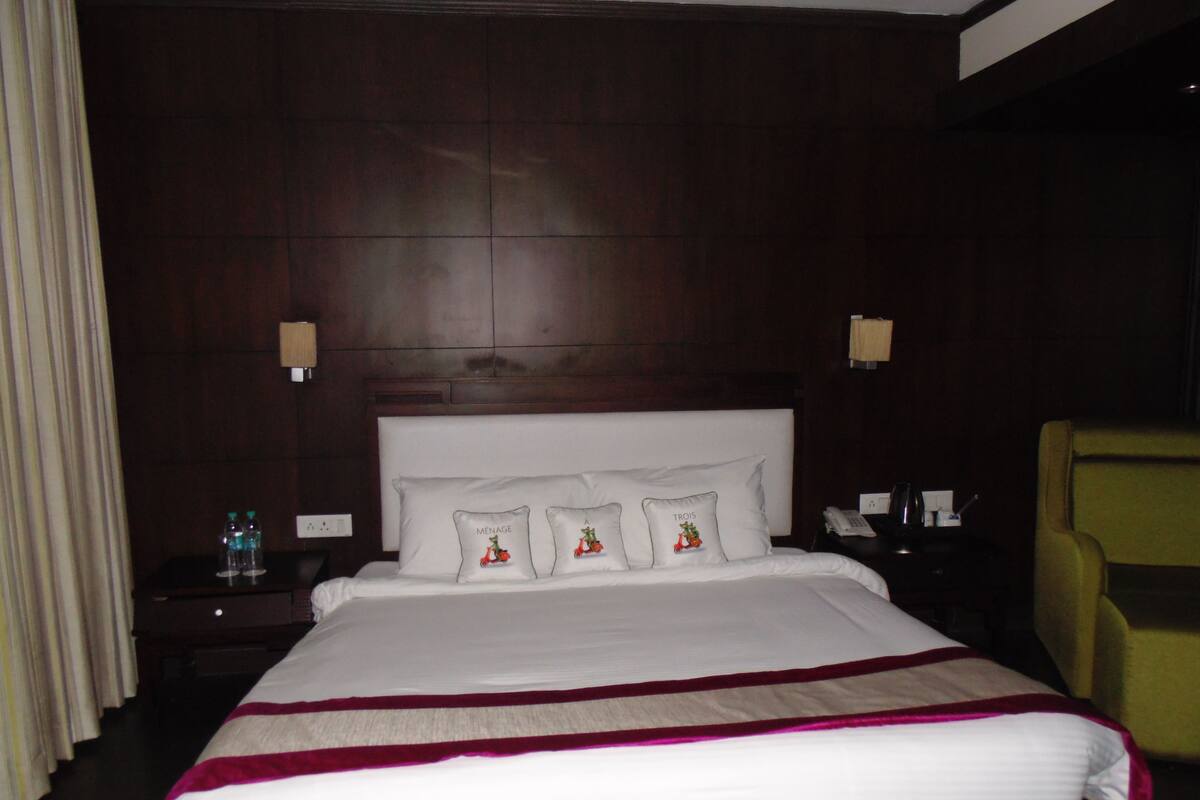 A hotel room with a large bed, a headboard with decorative pillows, a bedside table with a lamp, and a chair.