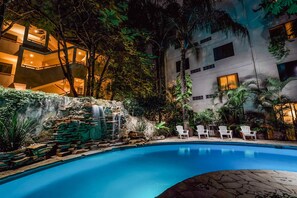 A swimming pool with a waterfall feature, surrounded by lush greenery and white chairs.