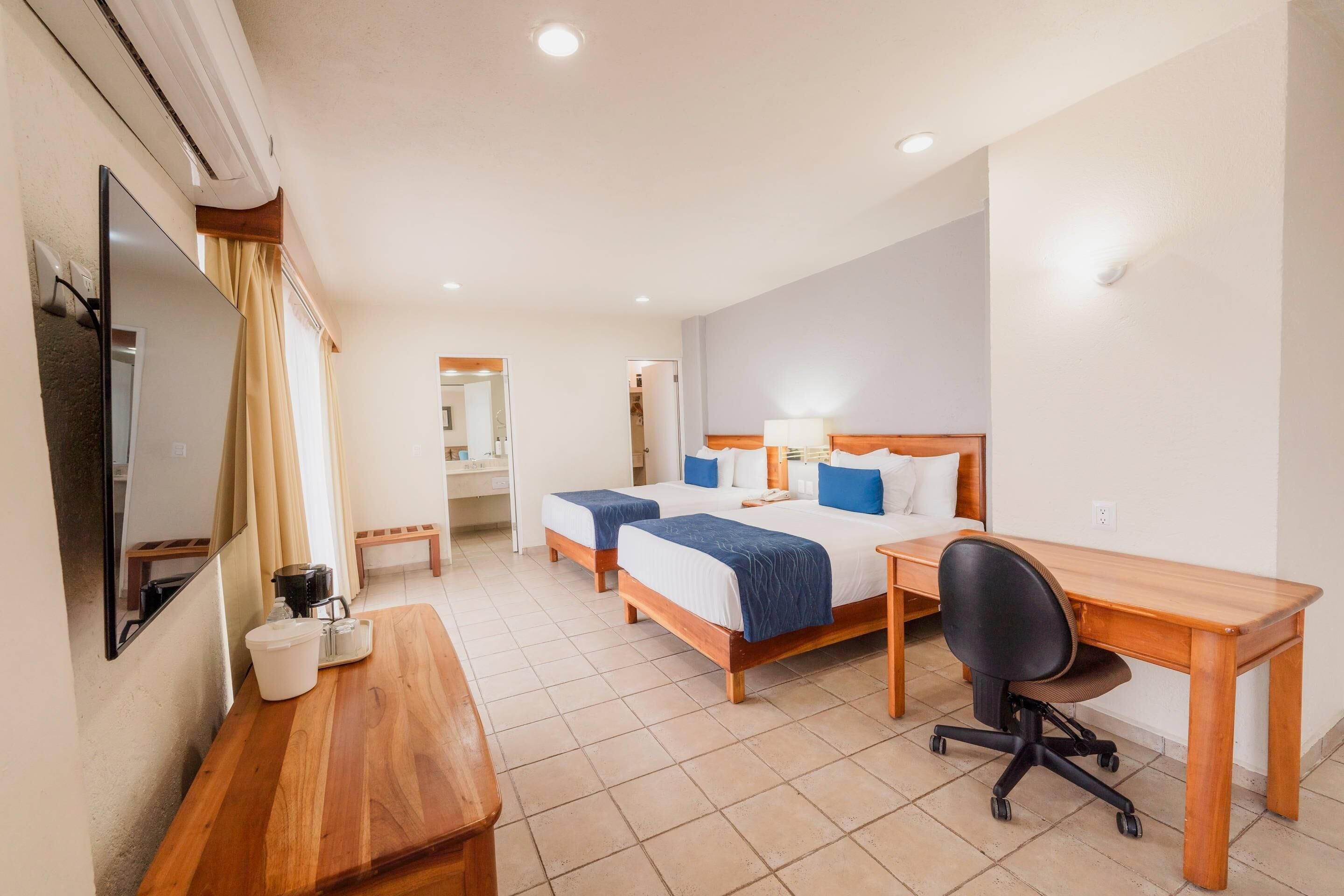A hotel room with two beds, a desk, a chair, a TV, and a bathroom visible in the background.