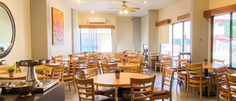 A spacious dining area with multiple wooden tables and chairs, ceiling fans, and large windows allowing natural light.