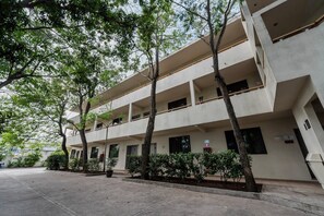 A multi-story building with balconies, a flat facade, and a paved ground area with trees and shrubs.