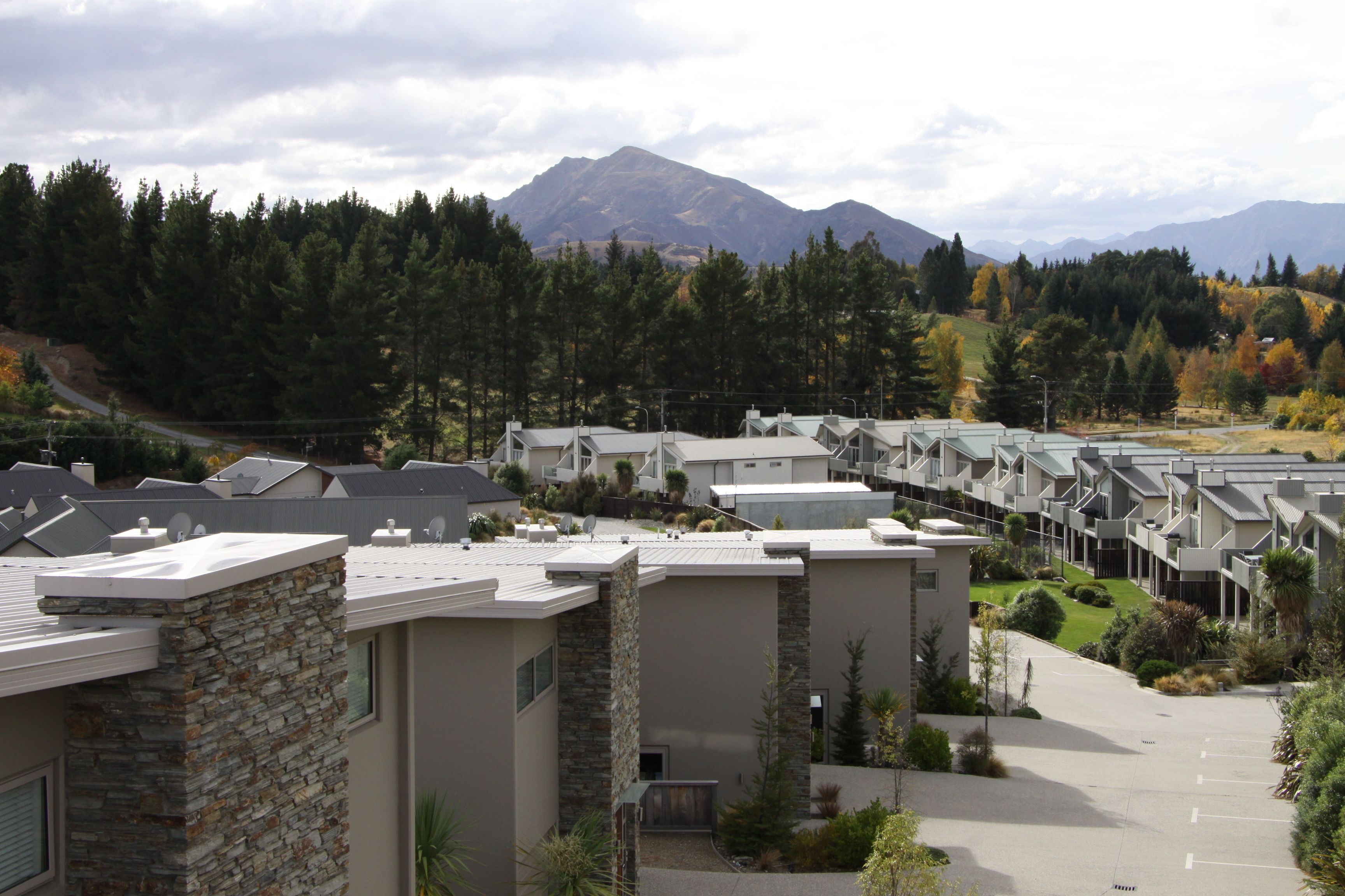 A row of modern buildings with stone facades, a parking lot, and a mountainous backdrop.