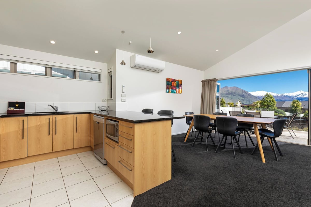 A modern kitchen with a large island, a dining area with a view of mountains, and a black carpeted floor.