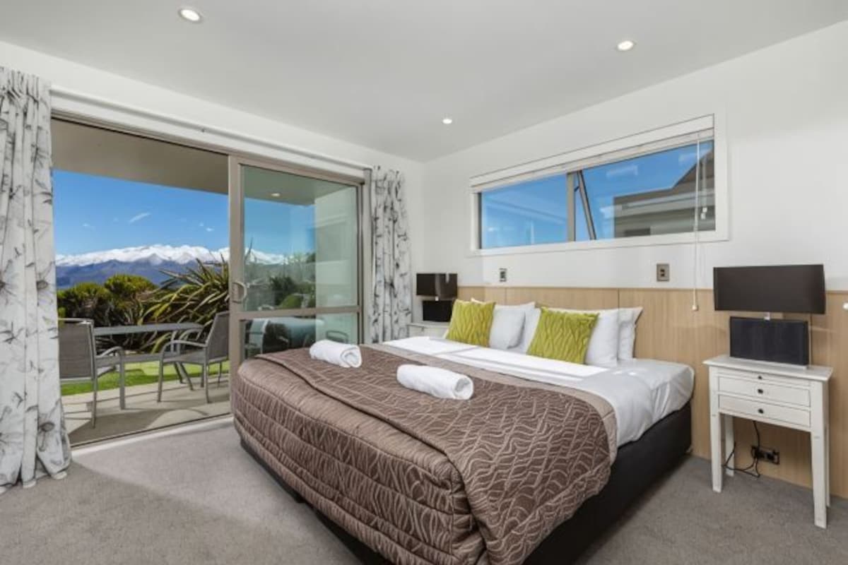 A hotel room with a bed, a TV, a desk, and a balcony with a mountain view.