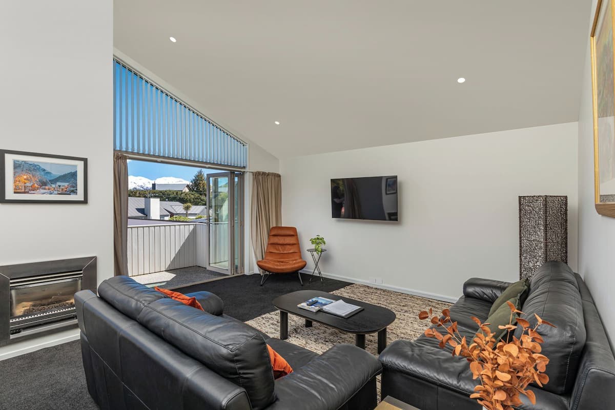 A modern living room with a sofa set, a coffee table, and a fireplace. There is a large window with a view of a mountain and a balcony.