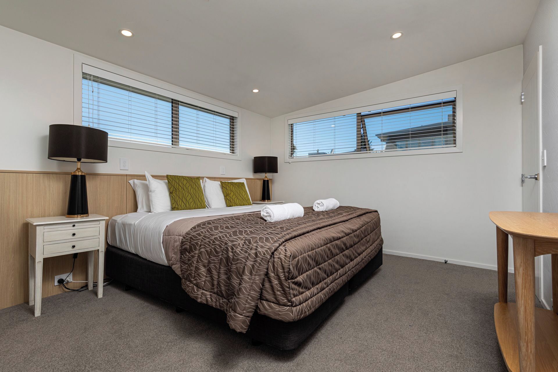 Une chambre moderne avec un grand lit, deux tables de chevet, une commode et deux fenêtres équipées de stores.