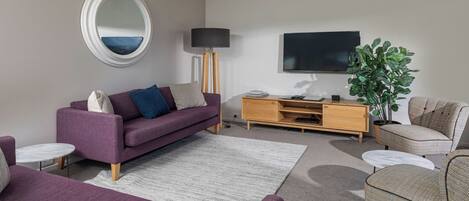 Un salon moderne avec un canapé violet, un miroir rond, un meuble TV en bois et une grande plante.