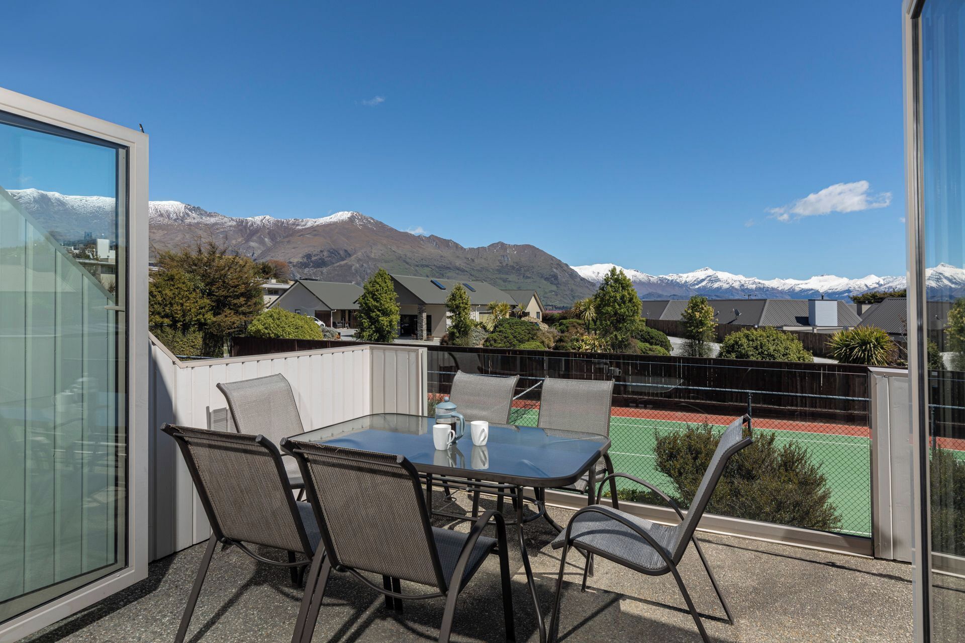 Un balcón con una mesa y sillas de vidrio, con vistas a una cancha de tenis y montañas.