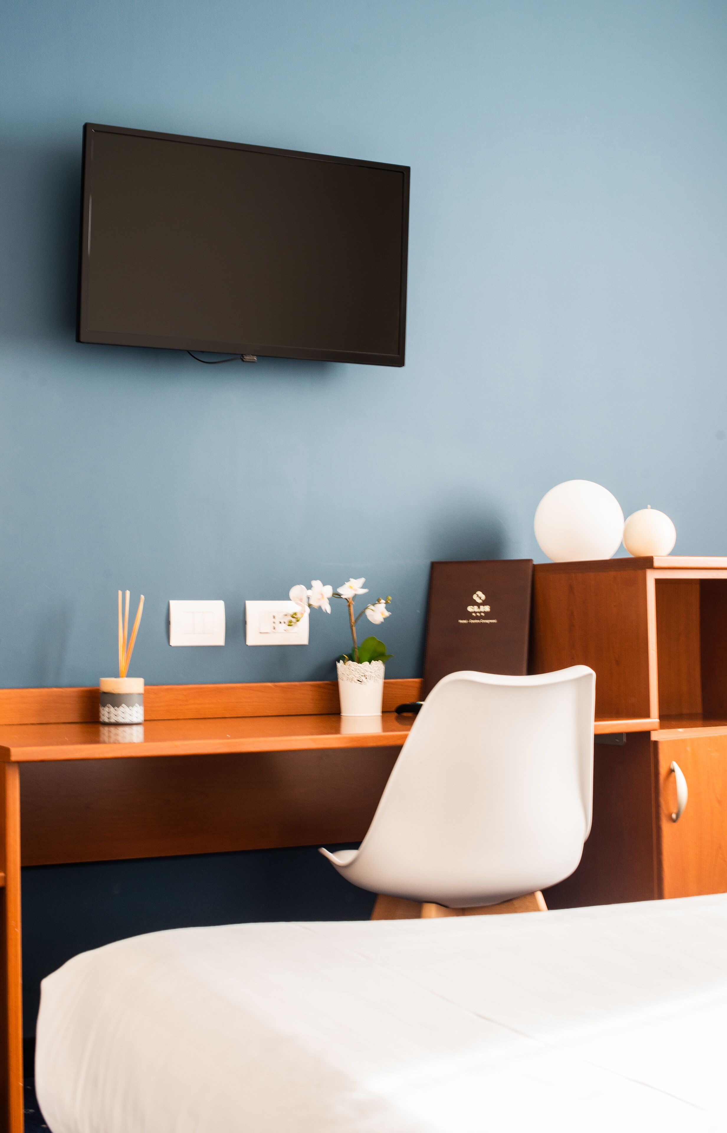 A hotel room with a bed, a desk with a chair, a television mounted on the wall, a small potted plant, and a decorative item on the desk.