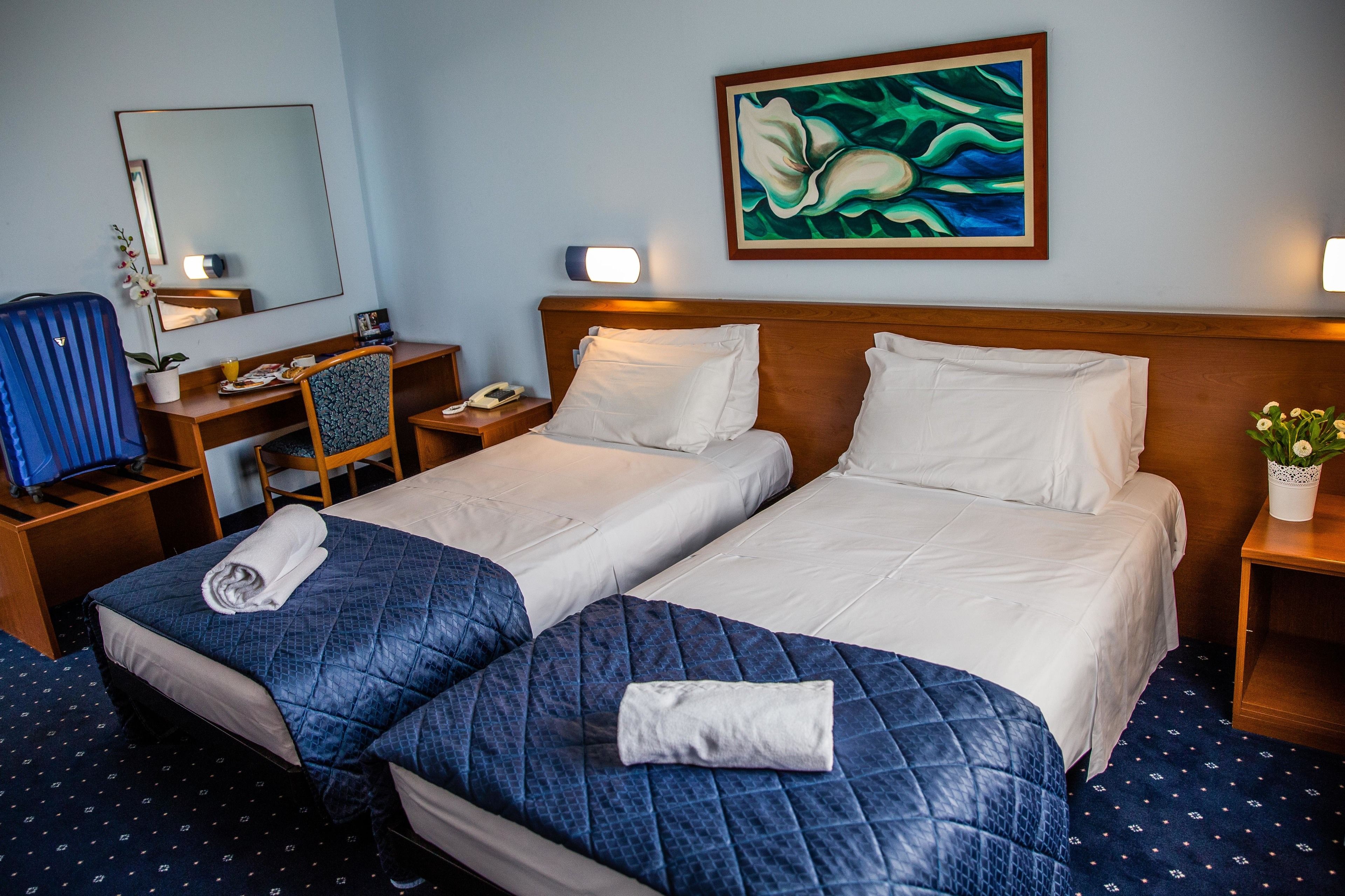 Habitación de hotel con dos camas, un escritorio, una silla, un espejo, un cuadro y una mesita de noche con lámpara.