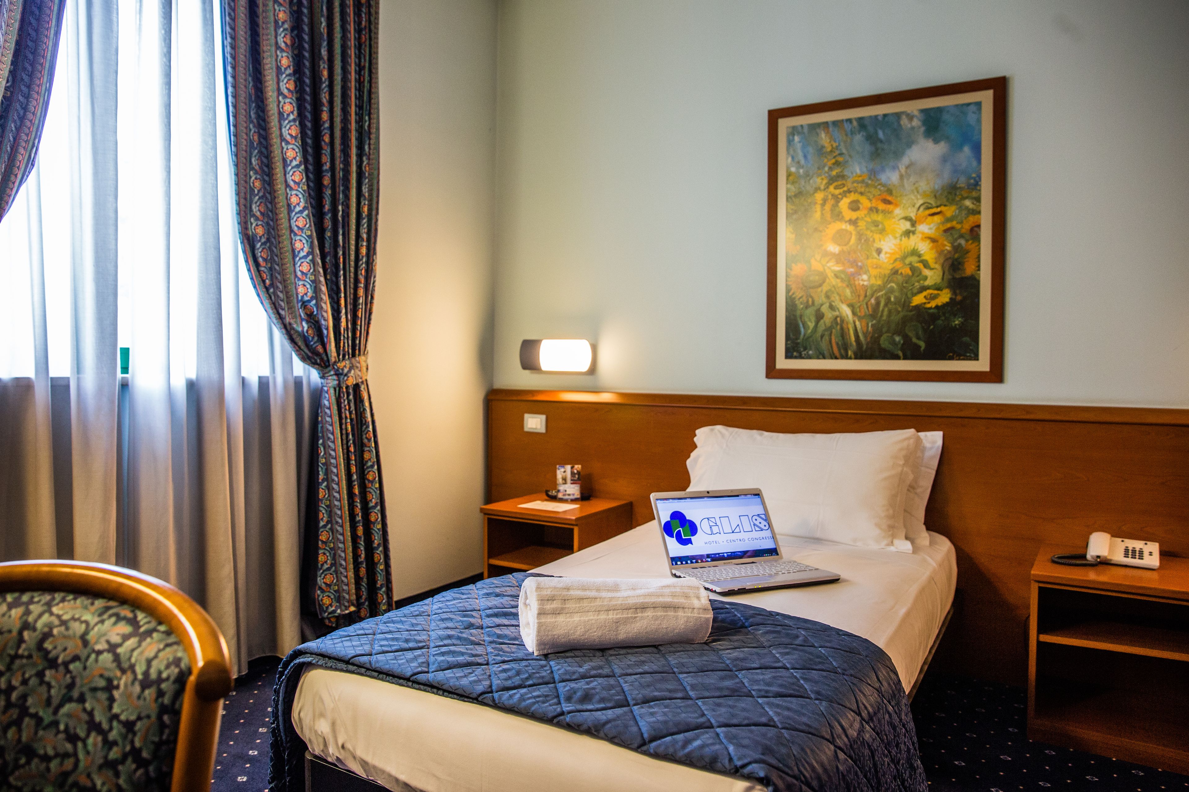 Habitación de hotel con cama, una laptop sobre la cama, un cuadro en la pared, una mesita de noche, una silla y cortinas.