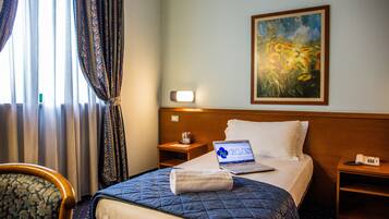 Habitación de hotel con cama, una laptop sobre la cama, un cuadro en la pared, una mesita de noche, una silla y cortinas.