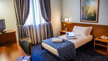 A hotel room with a bed, a desk, a chair, a laptop, a nightstand with a phone, and a painting on the wall.