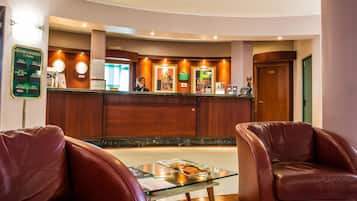 Una hall d'albergo con una reception, due poltrone in pelle, un tavolino in vetro e una segnaletica che indica la zona dei congressi e delle sale pianoforte.
