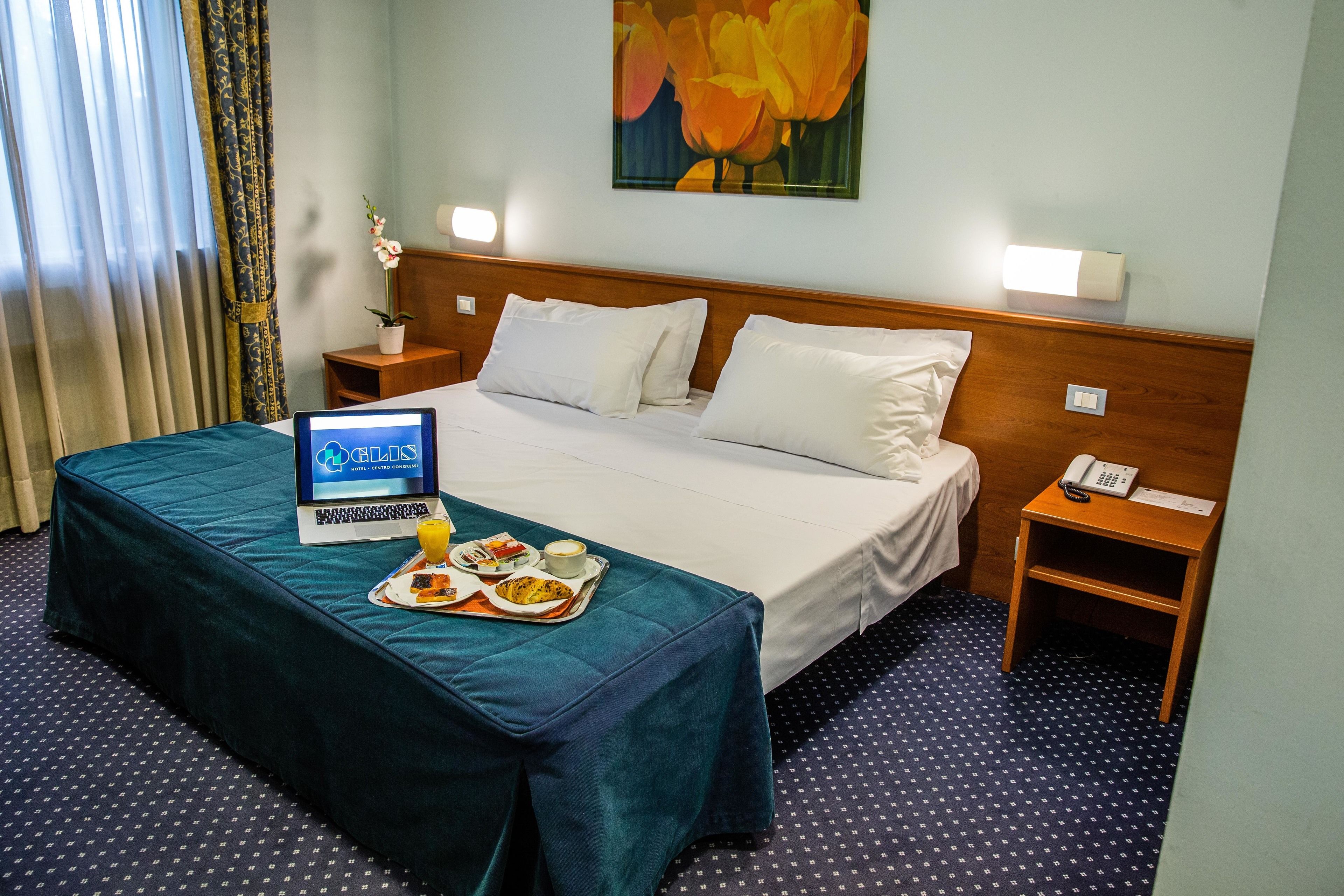 A hotel room with a bed, a laptop, a tray with food, and a painting on the wall.
