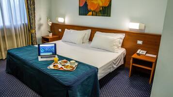 A hotel room with a bed, a laptop, a tray with food, and a painting on the wall.