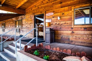 Une cabane en bois avec un escalier en bois menant à une porte portant l’inscription « BUREAU », un petit jardin délimité par une bordure en briques et une fenêtre avec des stores.