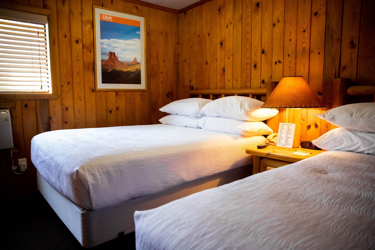 A room with two beds, wooden walls, a framed picture, and a lamp on a nightstand.