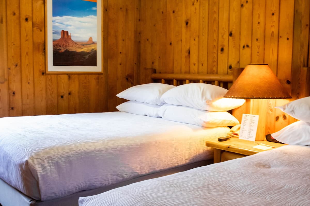 A room with wooden panel walls, two beds with white linens, a nightstand with a lamp, and a framed picture on the wall.