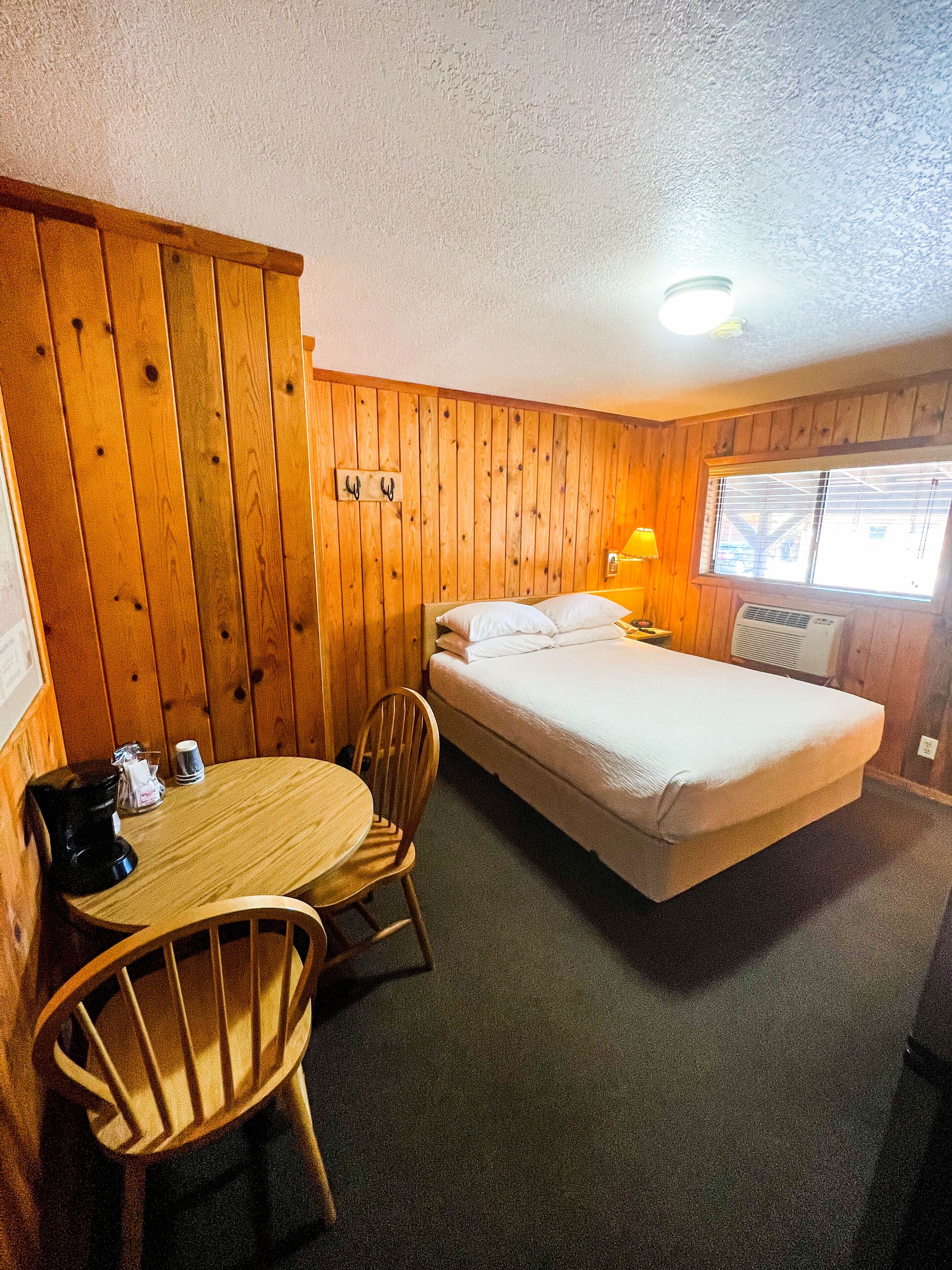 A room with a wooden paneled wall, a single bed with white bedding, a small round table with two chairs, a coffee maker, and a window with blinds.