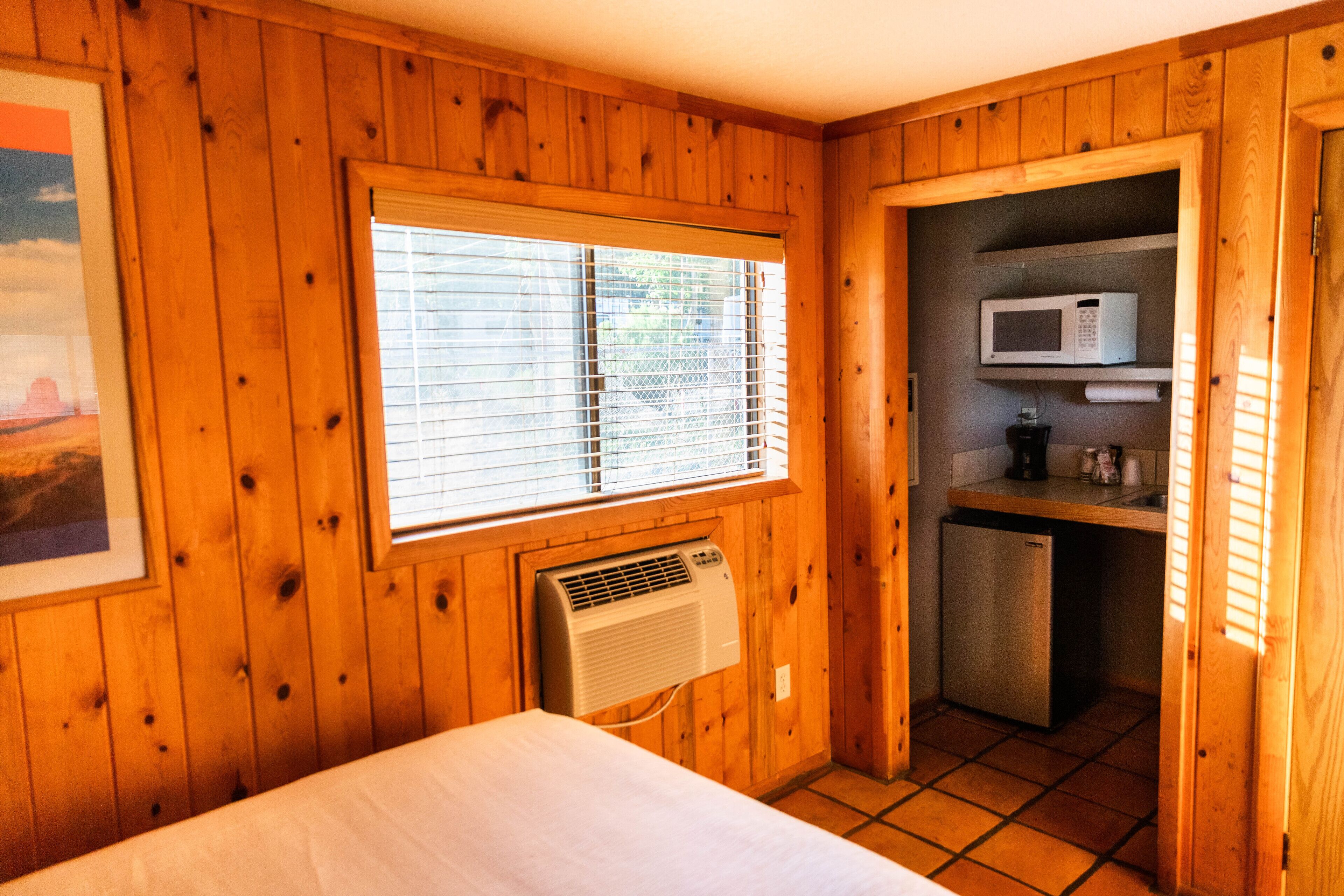 Una habitación con paneles de madera, una cama, una ventana con persianas, aire acondicionado integrado, un microondas y una pequeña cocina.