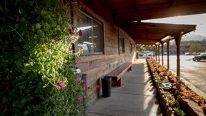 A covered outdoor area with a wooden bench, potted plants, and a view of a parking lot.
