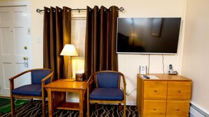 Habitación de hotel con dos sillones tapizados en azul, una mesa de madera con lámpara, un televisor montado en la pared y un aparador de madera con cajones.