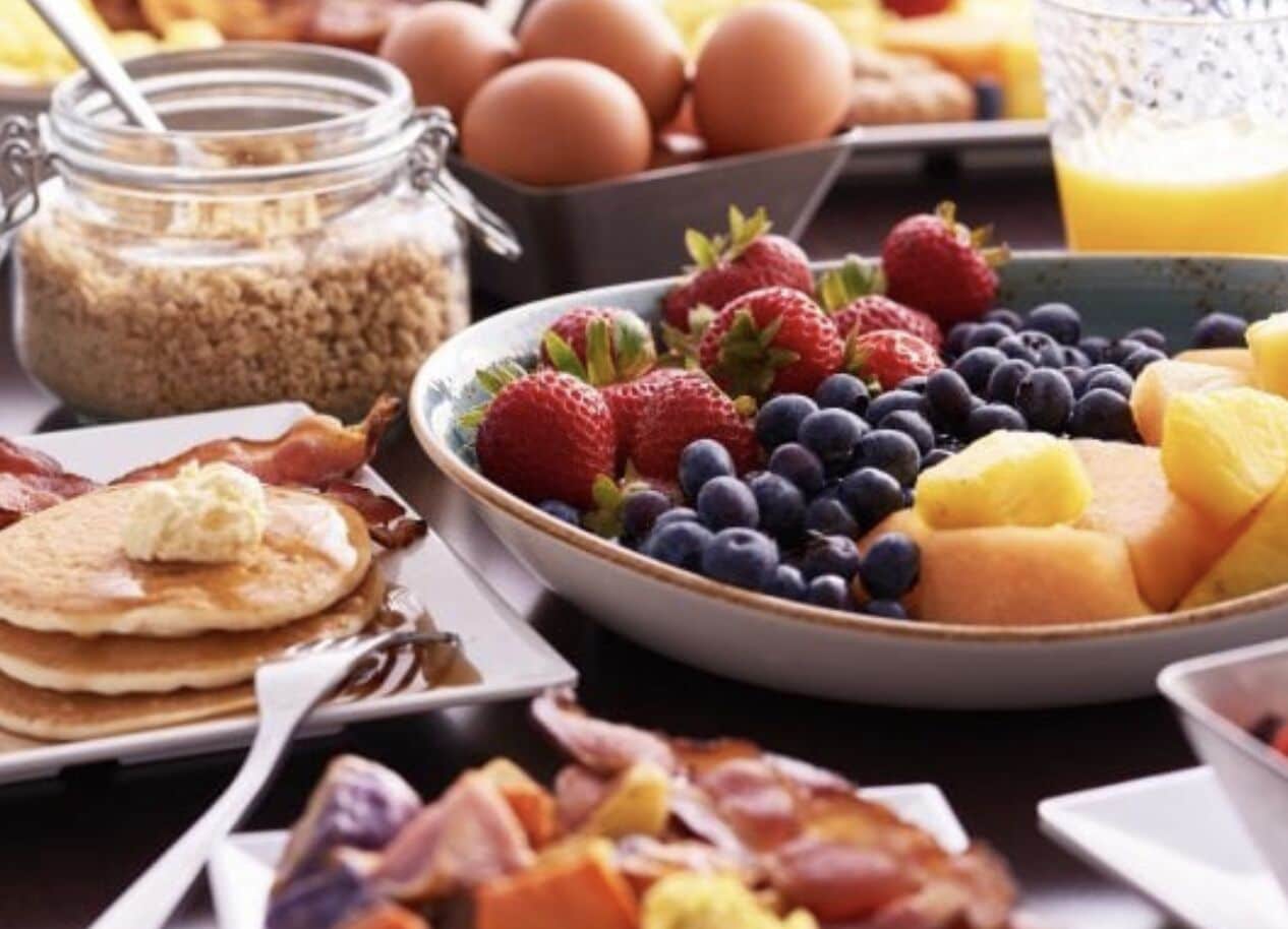 Une table de petit-déjeuner avec des crêpes, du bacon, des œufs et une variété de fruits, notamment des fraises, des myrtilles et des mangues.