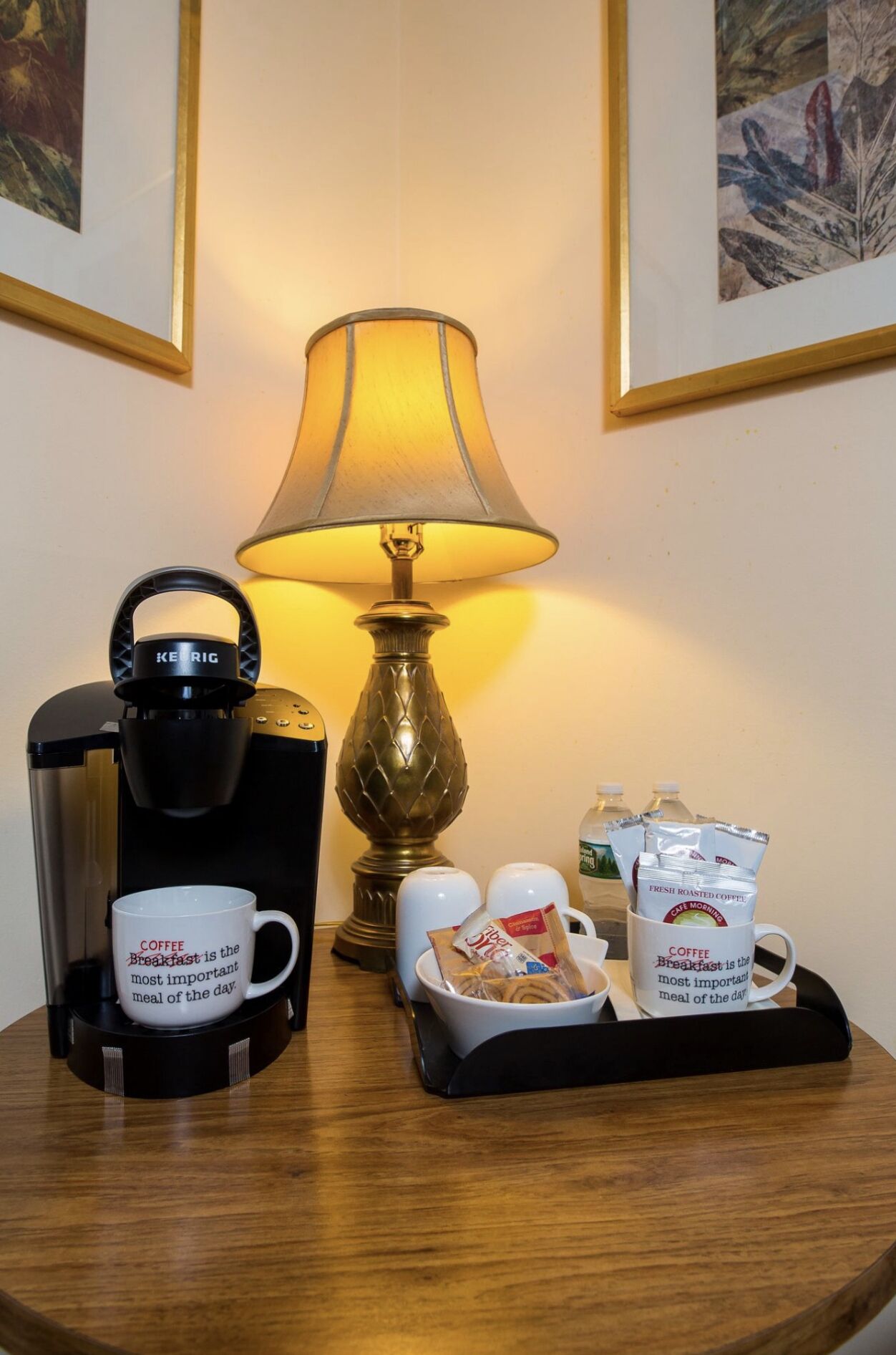 A coffee station with a Keurig coffee maker, two mugs, a tray with coffee packets, and a table lamp.
