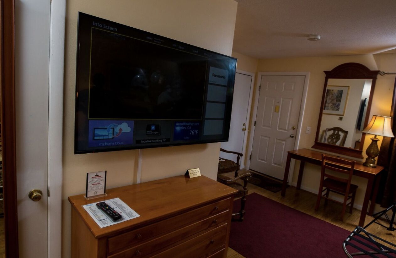 Habitación de hotel con una televisión de pantalla plana grande montada en la pared, un aparador de madera, un escritorio con lámpara y una silla.