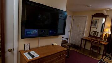 Habitación de hotel con una televisión de pantalla plana grande montada en la pared, un aparador de madera, un escritorio con lámpara y una silla.