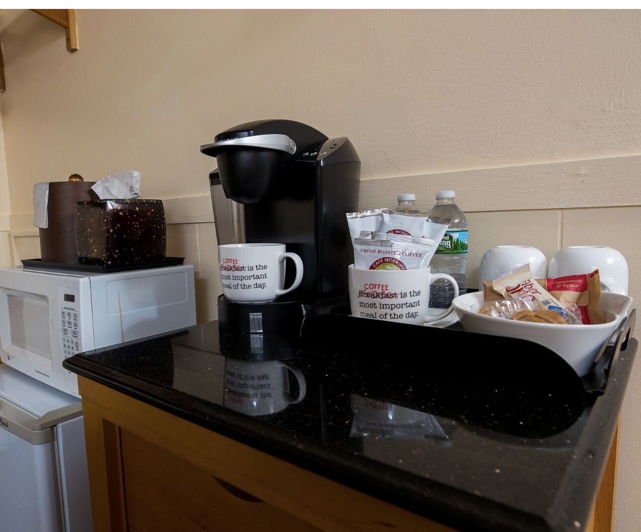 A kitchen counter with a coffee maker, two mugs with breakfast text, a water bottle, and a bowl of snacks.