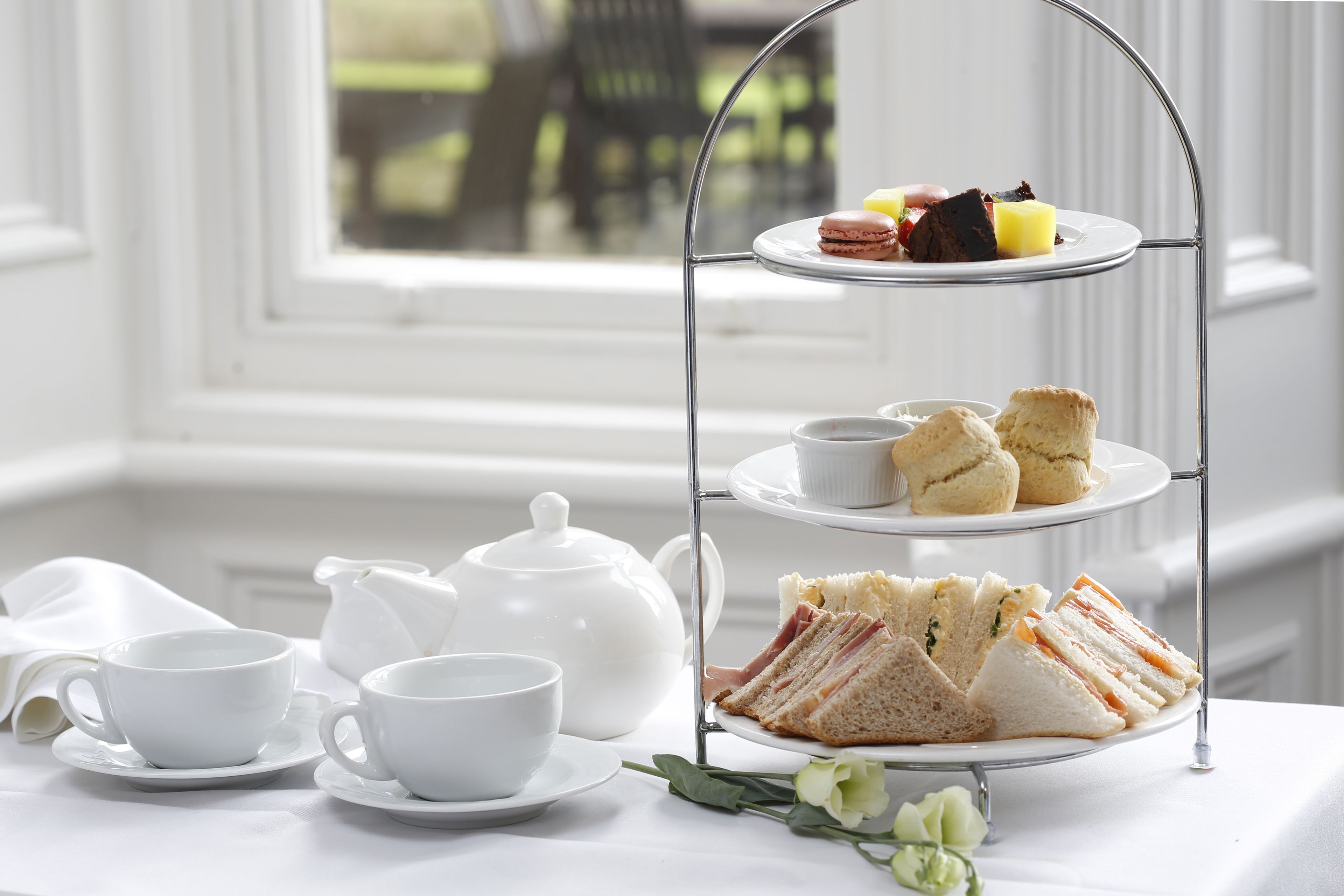 Un expositor de tres niveles para servicio de té, con sándwiches, scones, una tetera y tazas, sobre una mesa junto a una ventana.