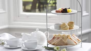Un expositor de tres niveles para servicio de té, con sándwiches, scones, una tetera y tazas, sobre una mesa junto a una ventana.