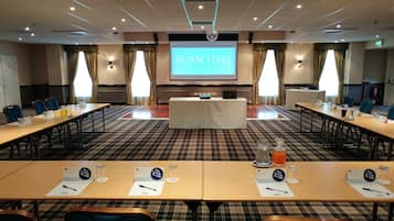 Ein Konferenzraum mit einer großen Leinwand, auf der “BURN HALL HOTEL, NEW YORK” steht, langen Tischen mit Namensschildern und einer Rezeption.