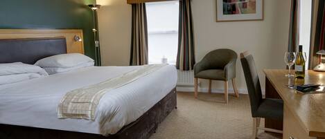 A hotel room with a large bed, a desk with a chair, a lamp, and a window with curtains.