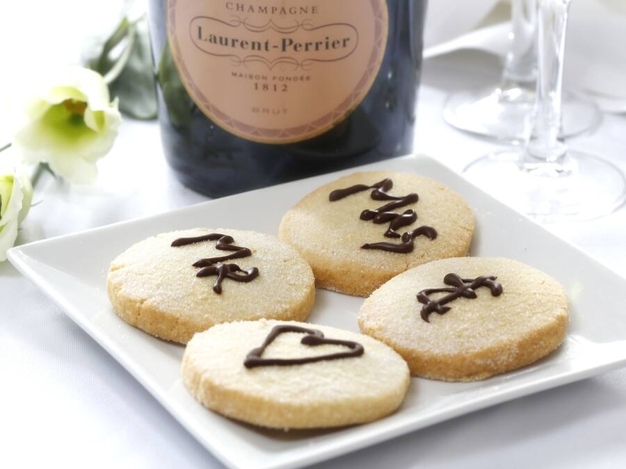 A plate of cookies with chocolate drizzle, a bottle of champagne, and a glass of wine on a table.