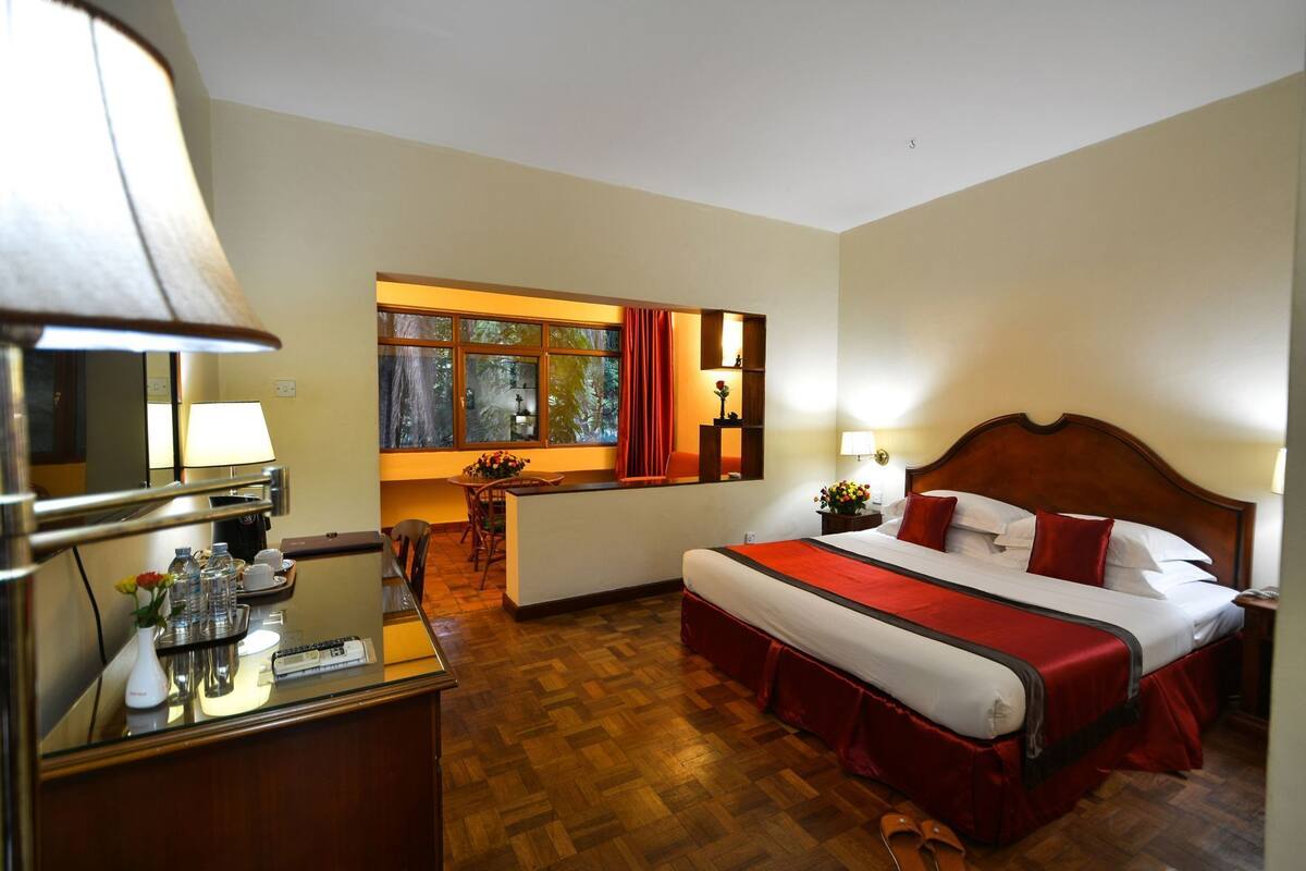 A hotel room with a bed, a desk with a lamp, a chair, a small table with a vase, and a window with curtains.