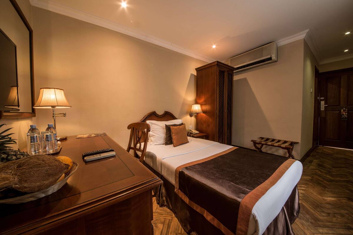 A hotel room with a bed, bedside table, lamp, and a small wooden bench.