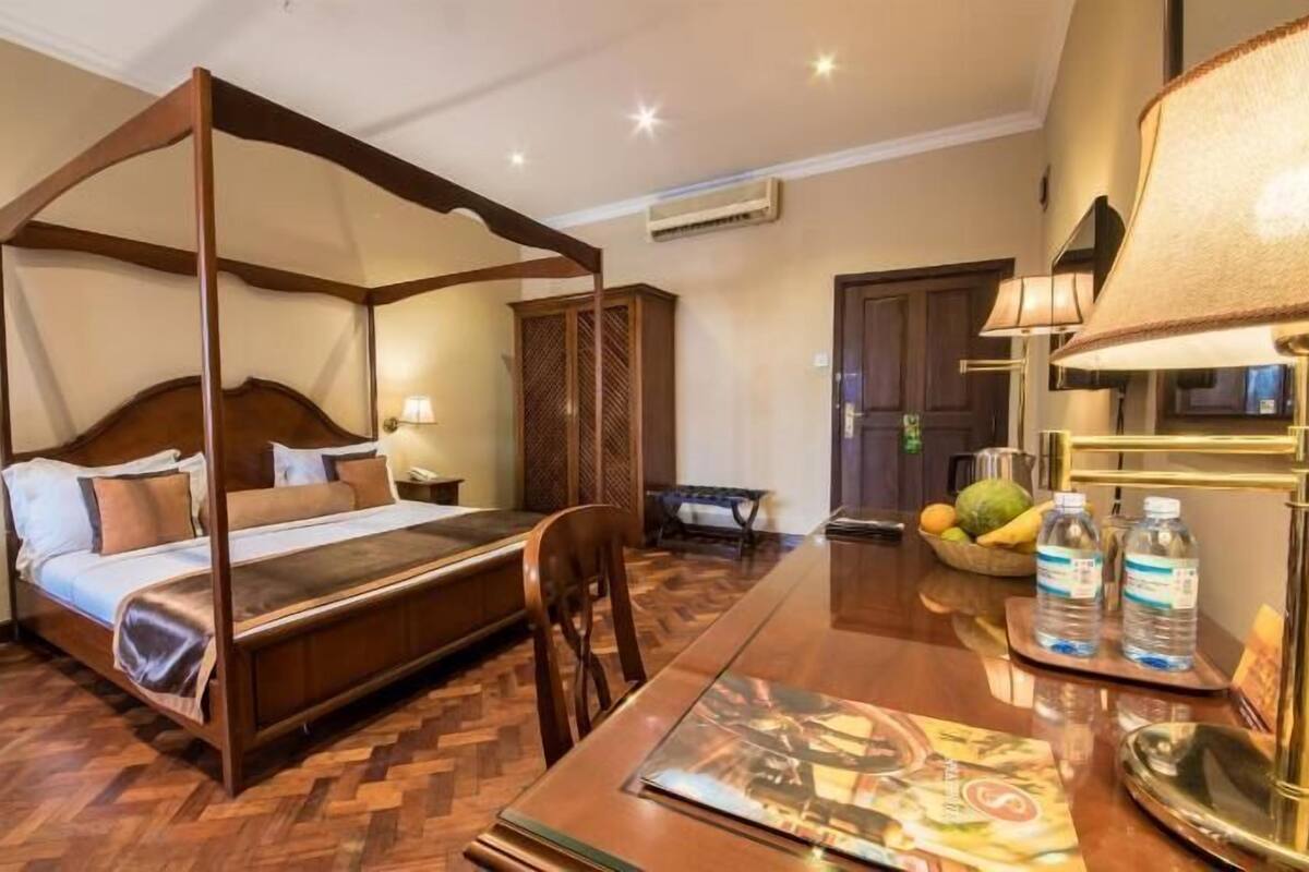 A four-poster bed with a wooden headboard, a dining table with chairs, and a fruit bowl on the table.