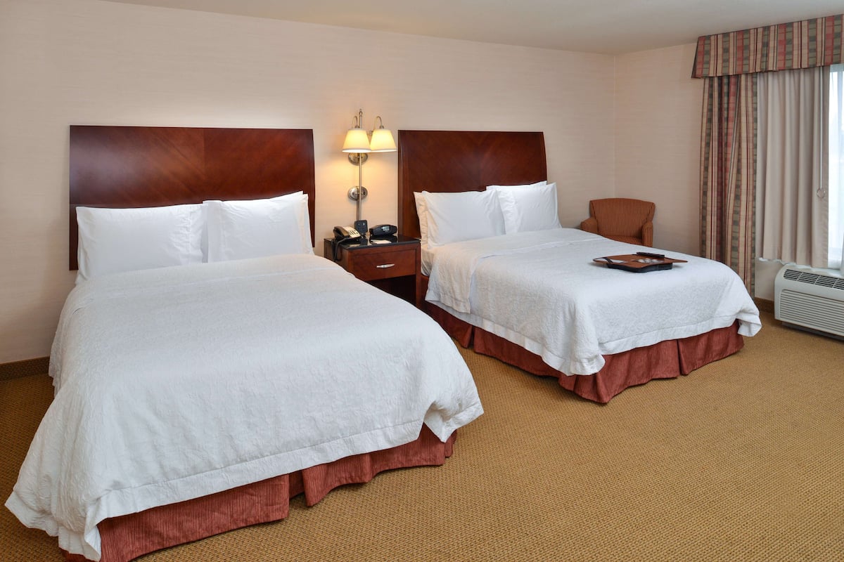 A hotel room with two beds, a nightstand with a lamp, a chair, and a window with curtains.