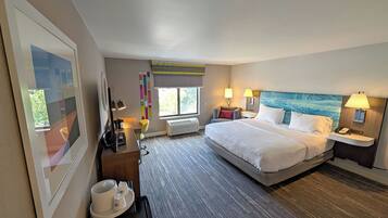 A hotel room with a large bed, bedside tables, a desk with a chair, a window, and a view of greenery outside.